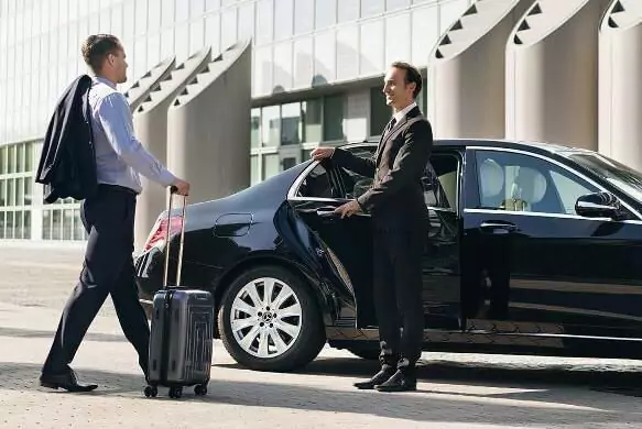 A professional chauffeur opening the door of a luxury car for a business traveler with luggage.