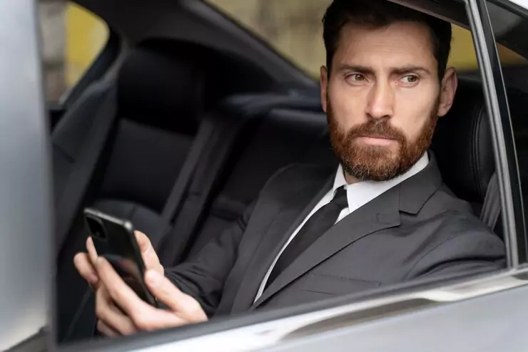 A well-dressed businessman with a beard sitting in the backseat of a luxury taxi, holding a smartphone and looking outside.
